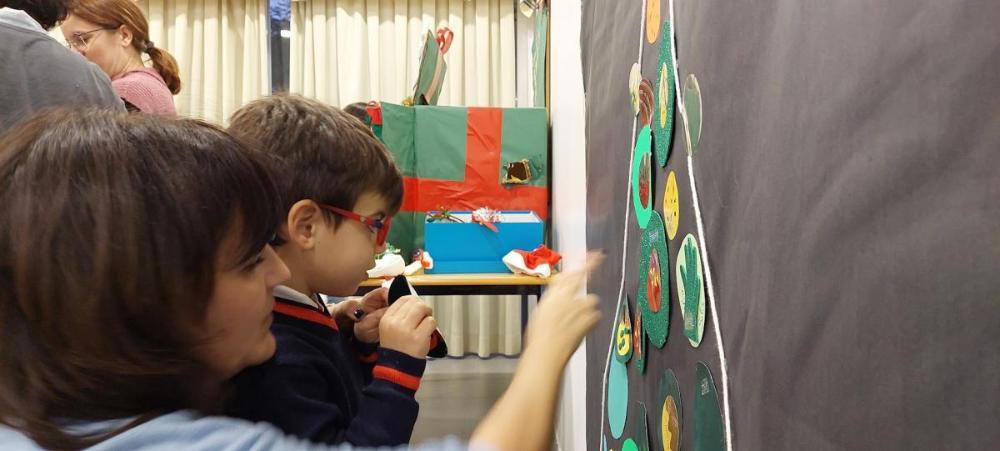 Maestra y alumno colocan su manualidad en el árbol de  navidad que está pegado a  la pared